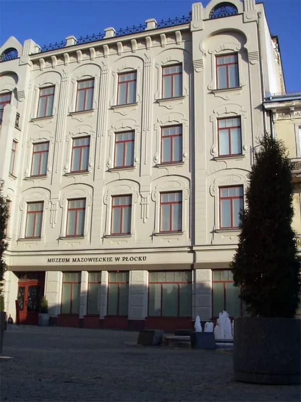A grand, light-colored building with multiple floors of windows, a decorative roofline, and a sign identifying it as "MUZEUM MAZOWIECKIE W PŁOCKU" above its entrance.