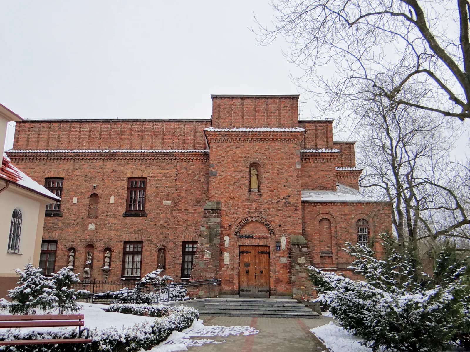 A historic brick building with a large wooden door and multiple statues embedded in the wall, surrounded by snow-dusted grounds and bare winter trees.