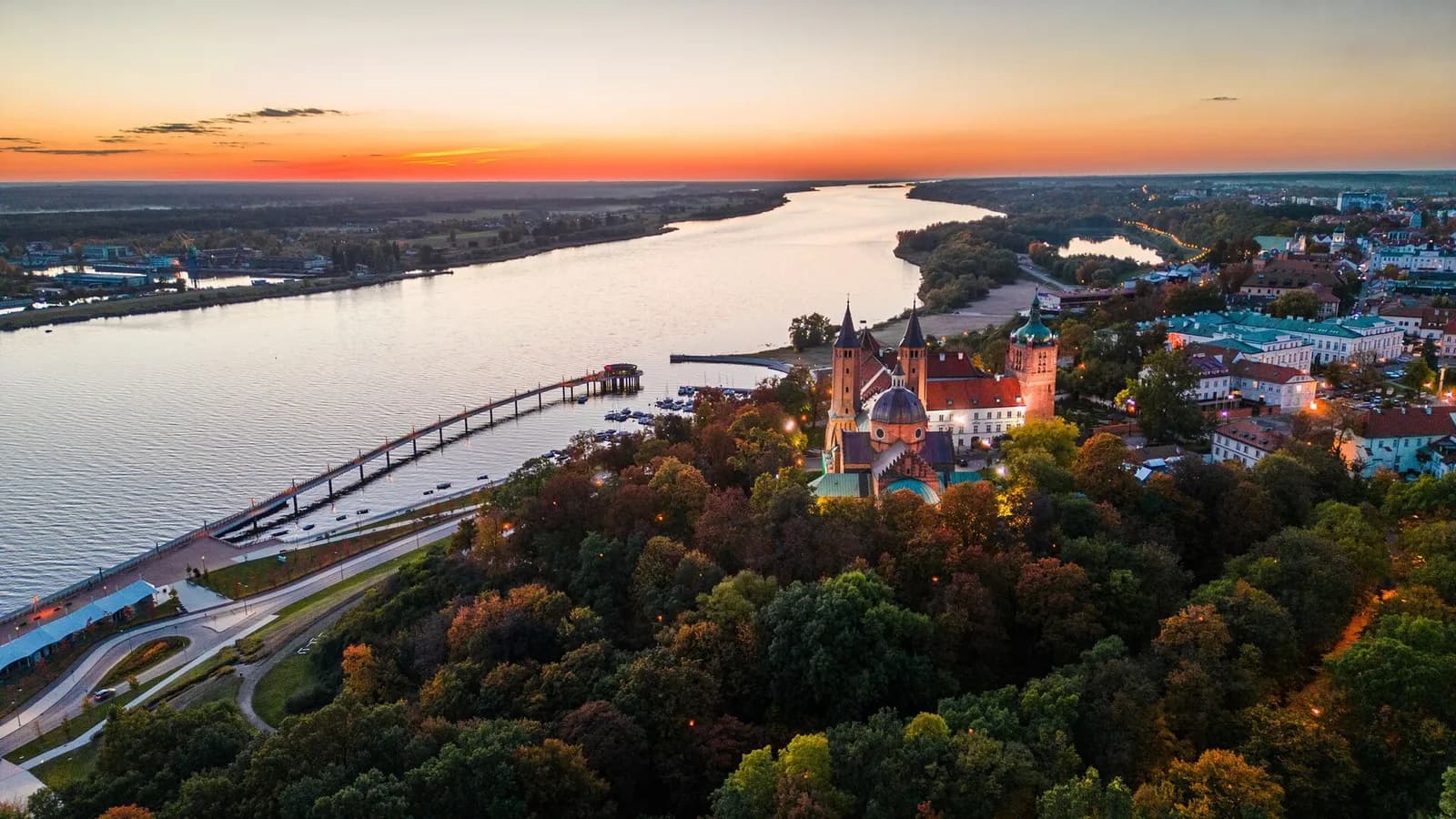 An aerial view at sunset shows a large river alongside a town with a prominent castle or cathedral nestled among trees on a hill.